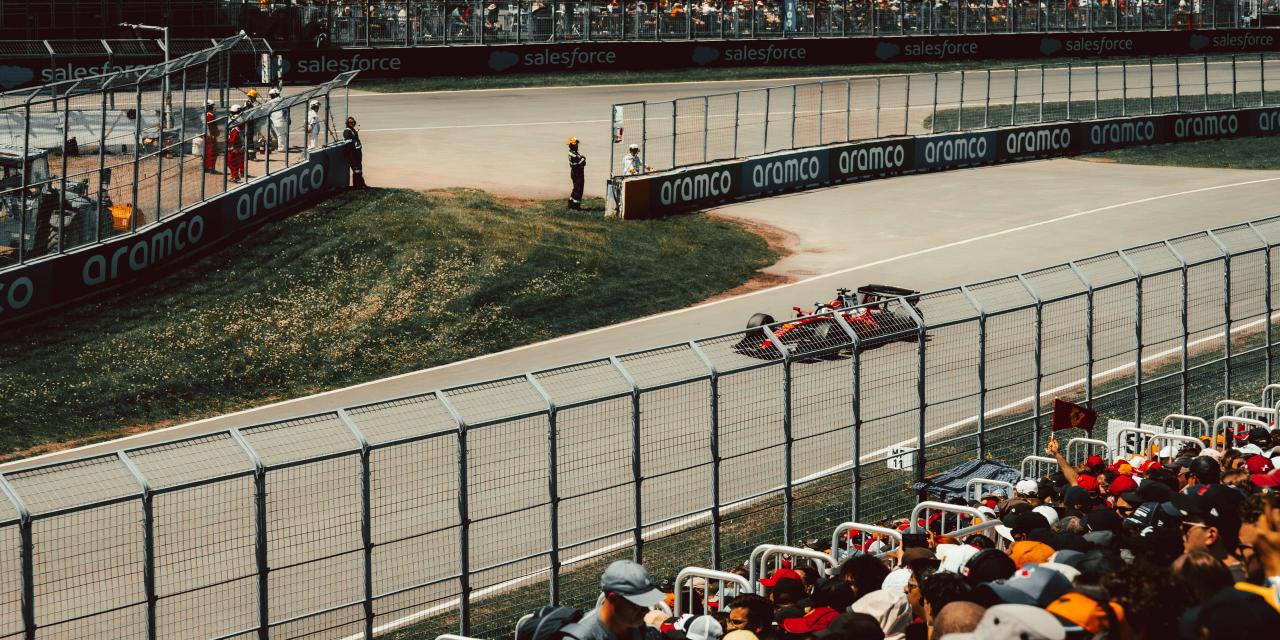 A Ferrari turning away into a corner, seen over the heads of a packed grandstand of spectators in caps, one of them holding up a Ferrari flag.