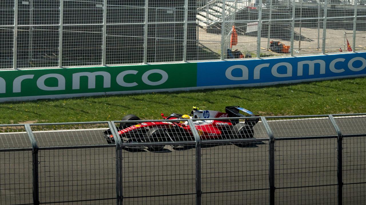 A red Ferrari Formula One car at speed along the barrier, shot through two layers of catch fencing, with sponsor boards and a marshal post running behind it.