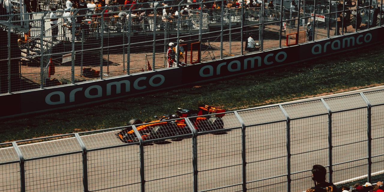 A papaya-orange McLaren Formula One car passing the barrier at speed, with spectators standing behind the fencing on the far side of the track and a marshal at his post.